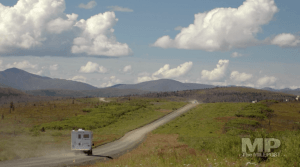 Truck with cab-over camper drives a gravel road mountains in background