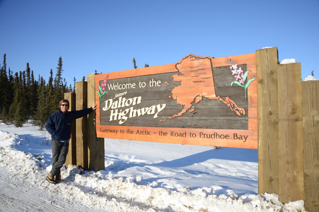 Traveler poses by a sign that says Welcome to the Dalton Highway and has an outline of Alaska