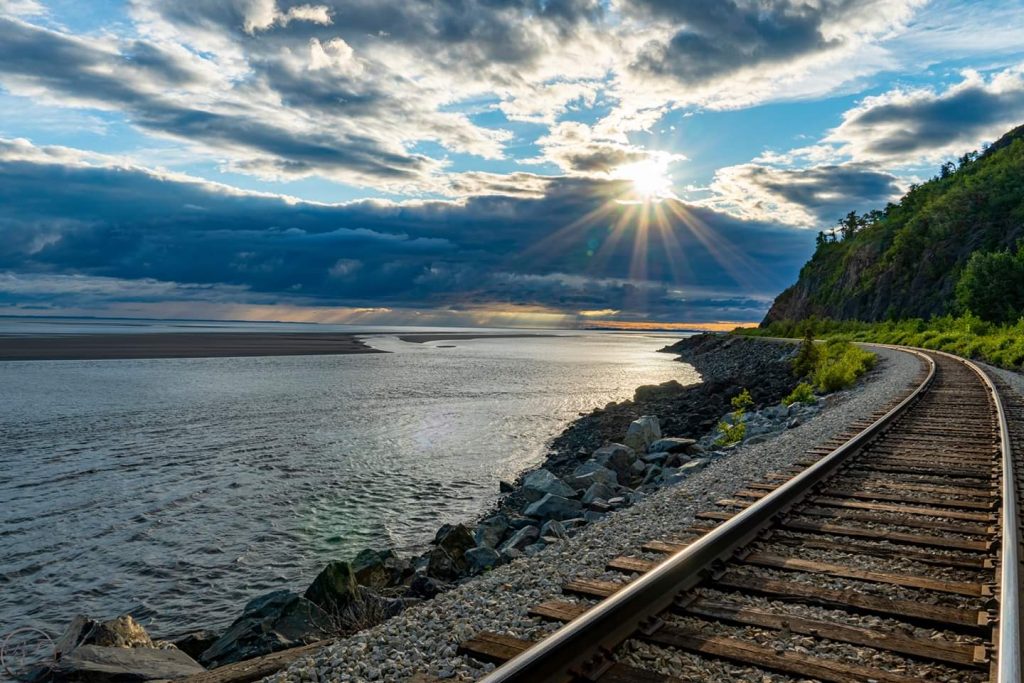 Sun sets behind clouds over water and winding train tracks on the shore