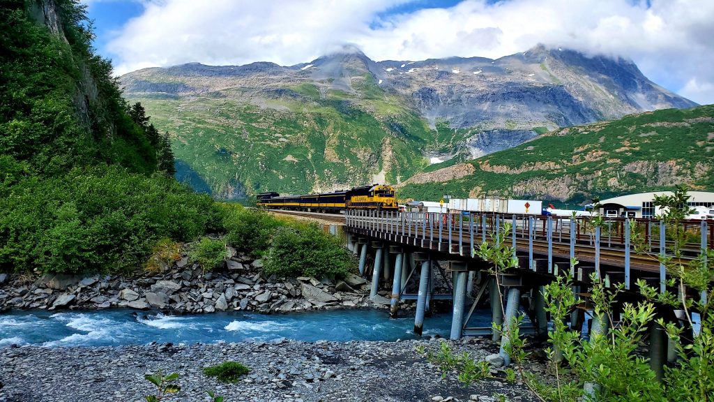 Alaska Railroad train crossing a bridge over a river with scenic mountains behind