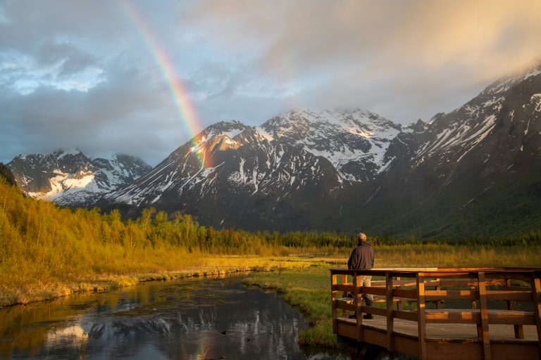 Man stands on wooden deck and looks out over valley, river, mountains, and a rainbow