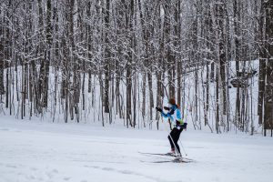 a red-haired woman cross country skiing along trail next to a stand of leafless trees