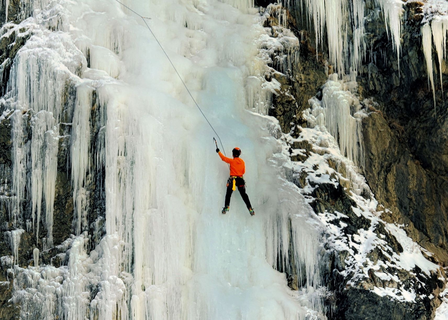 ice climber in orange jacket climbing up frozen waterfall