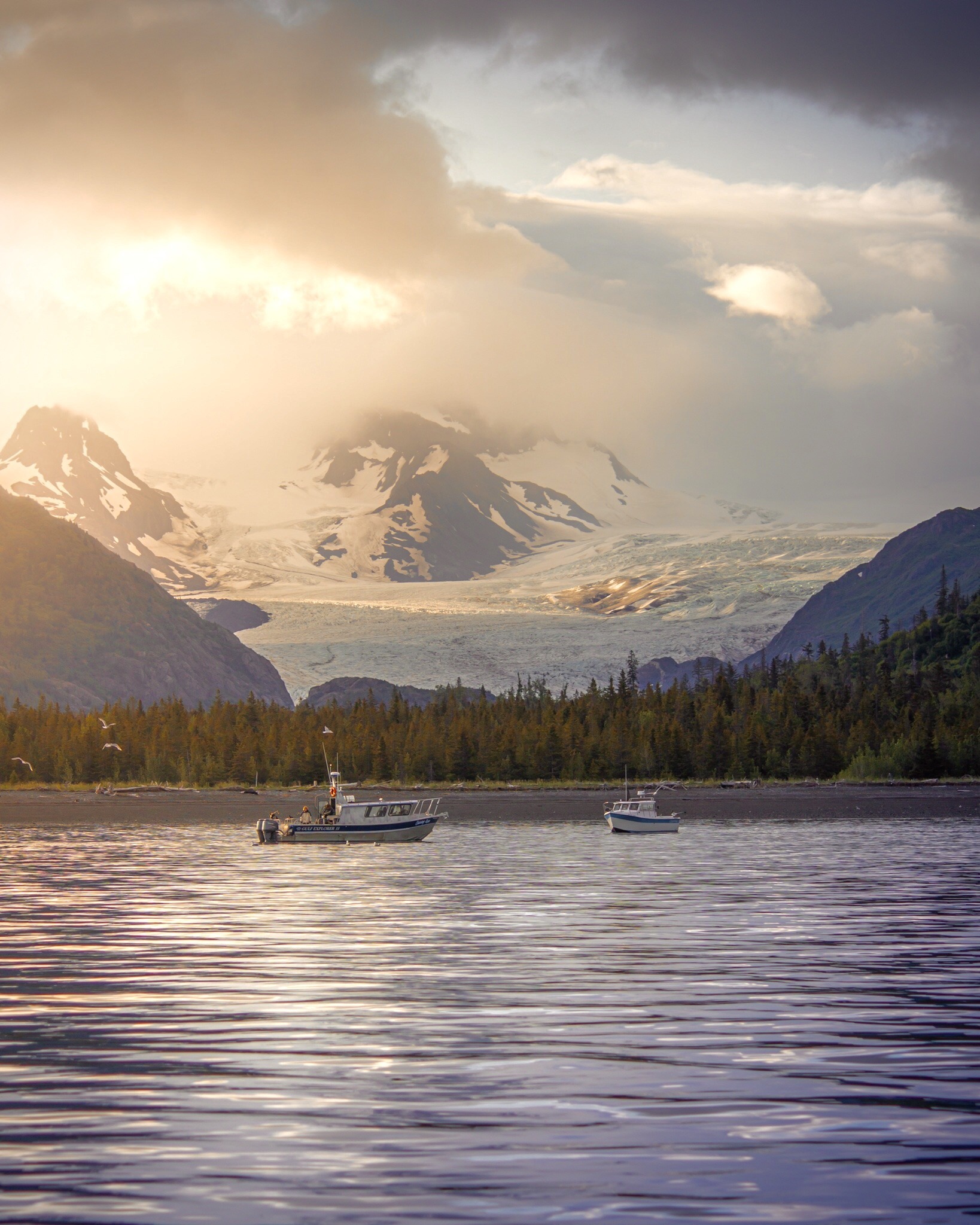Fishing boats on water, sun shining through clouds, glacier descending from mountains