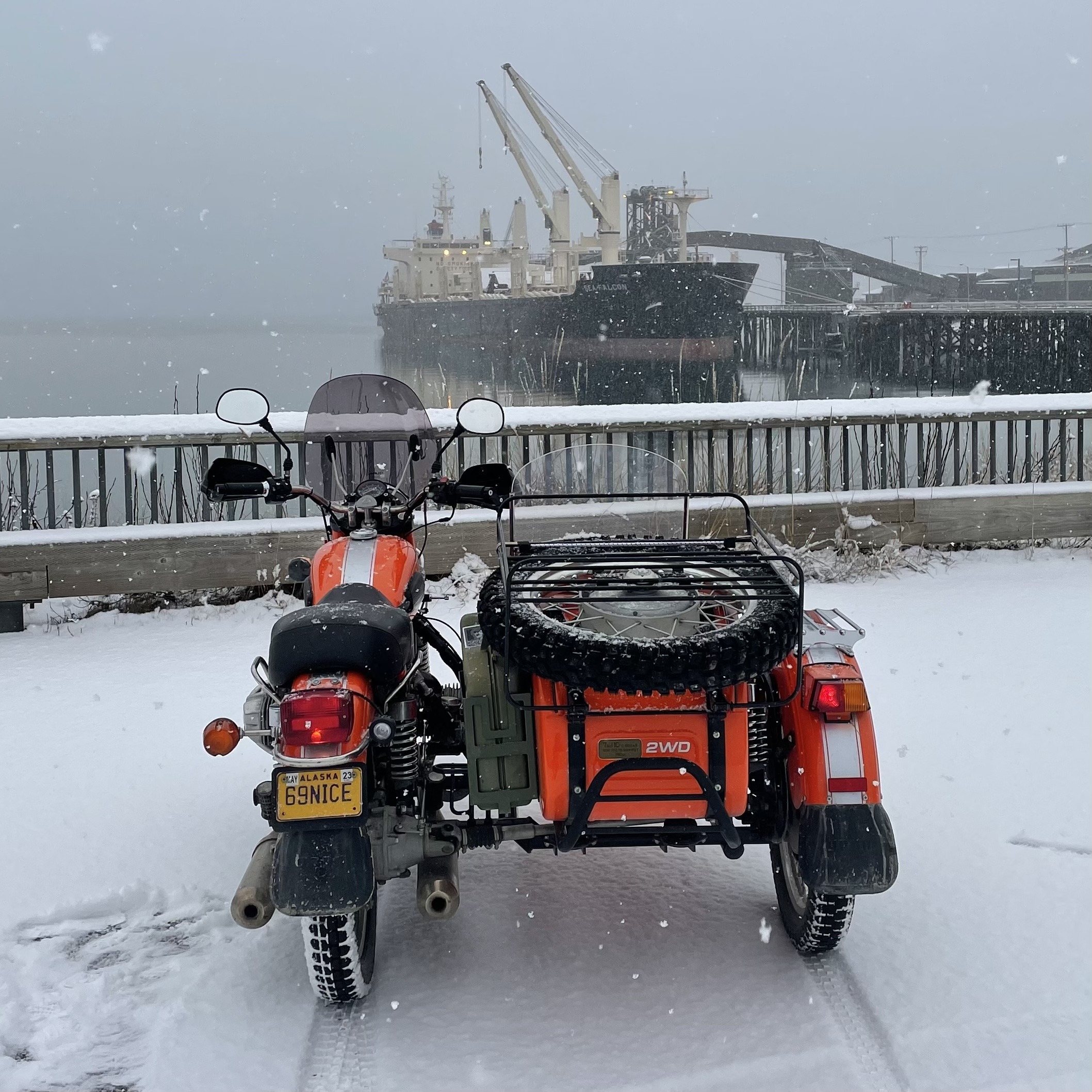 Orange motorcycle and side car parked in snow, ore ship being loaded on docks behind, snow falling