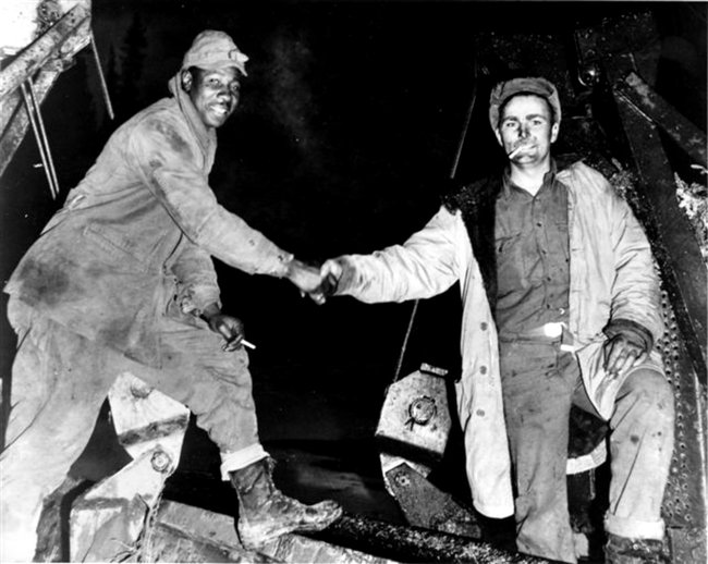 A black man and a white man, both dressed warmly, pose for the camera and smile while shaking hands