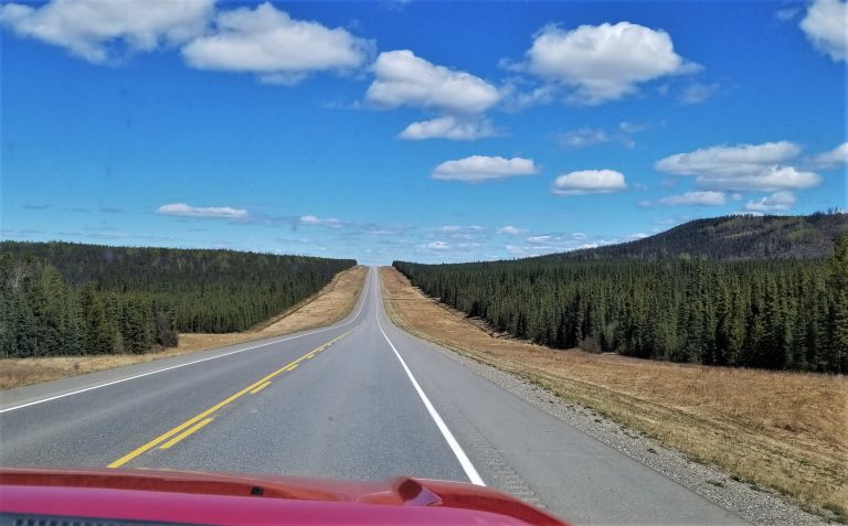 A straight stretch of the Alaska Highway stretches ahead past a red truck hood