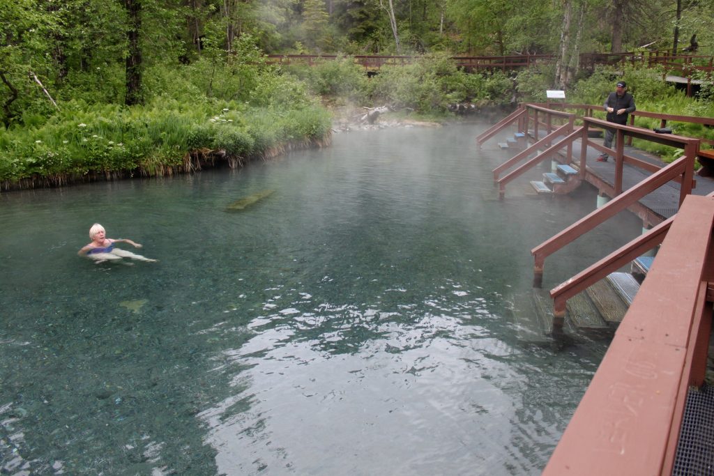 People soak in shallow hot spring next to a wooden platform and surrounded by forest greenery