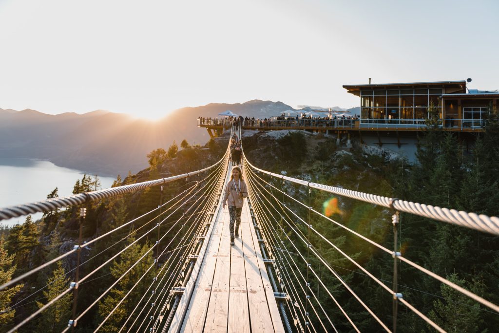 Sun sets behind mountains and a suspension bridges stretches between peaks. Summit lodge with dining deck is at the far end of the bridge.